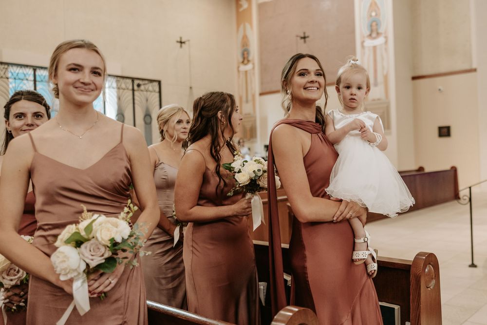 Bridesmaids in dusty rose dresses holding bouquets stand in church during wedding ceremony.
