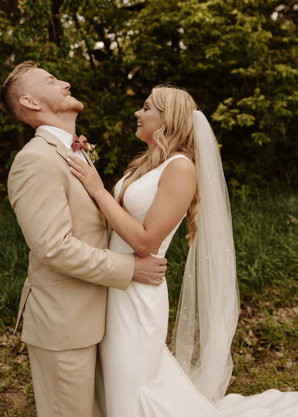 Romantic wedding couple laughing and embracing during a photo shoot in a natural outdoor setting.
