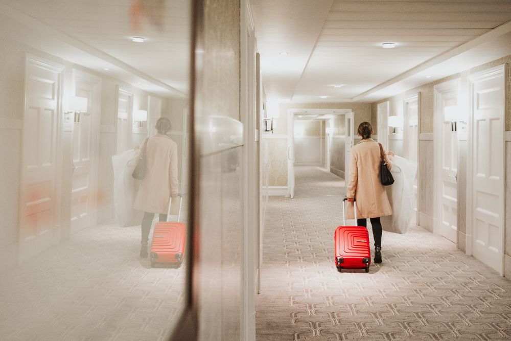 Person in beige coat walking down hotel hallway with red suitcase reflected in mirror.