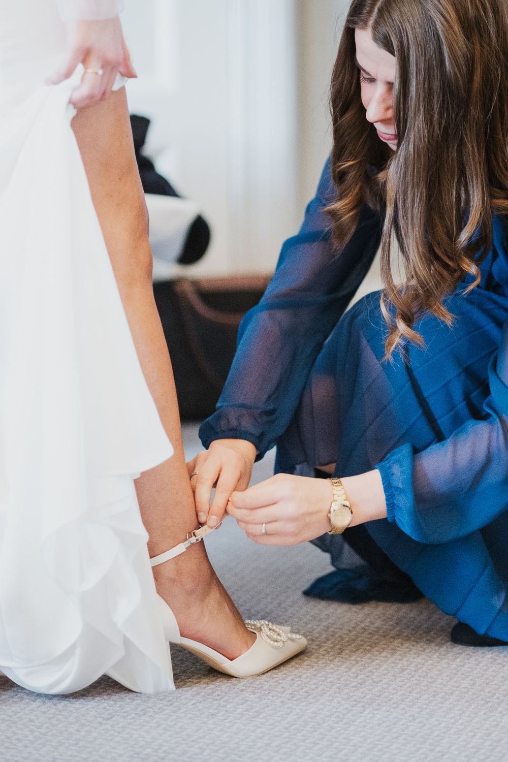 A person in a royal blue dress helps adjust the ankle strap on a bride's white flat wedding shoes.
