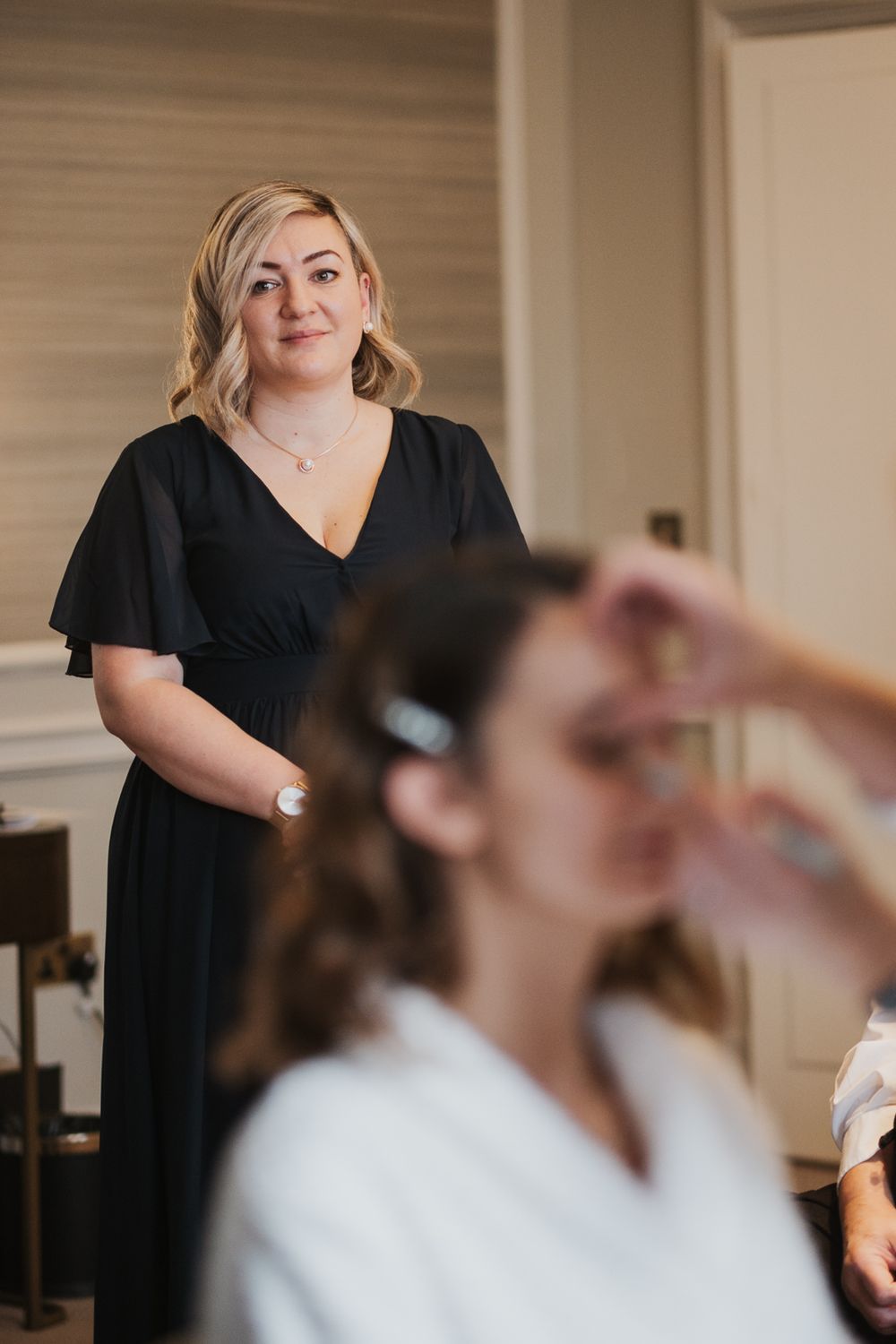 Woman in black dress stands watching while someone gets makeup applied in a bridal preparation setting at intimate london wedding.