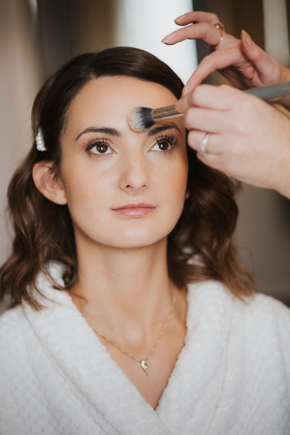 Professional makeup artist applying eyeshadow with a brush while client wears white robe and delicate necklace at an intimate london wedding