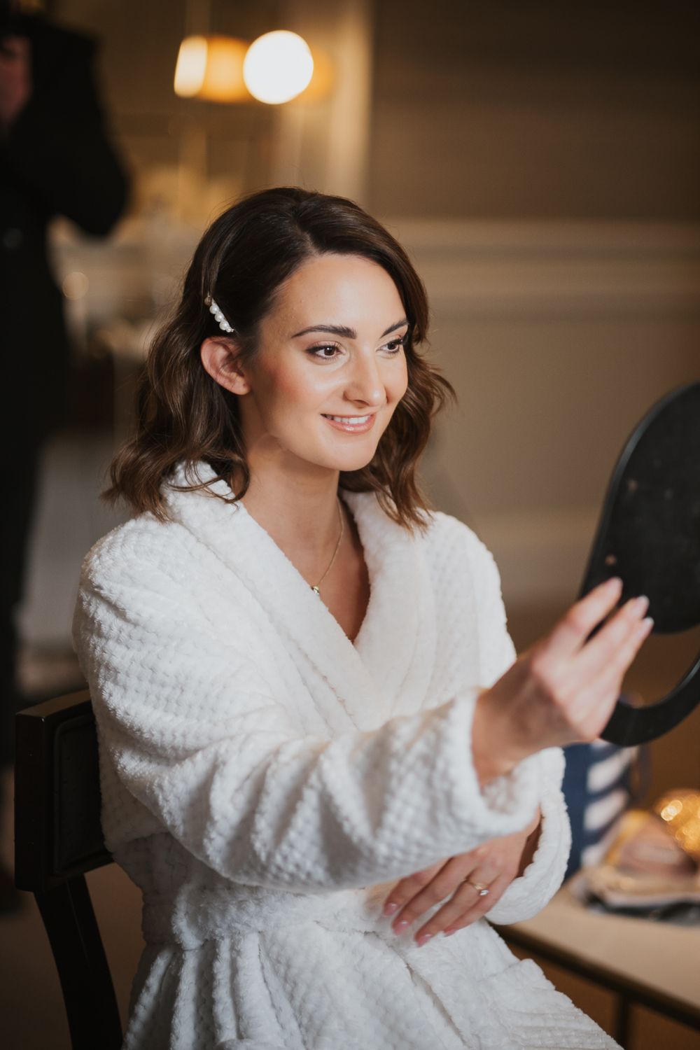 A woman in a white bathrobe smiles while looking at herself in a mirror during preparation.