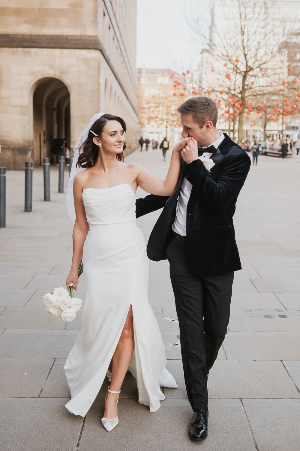 A newlywed couple walking on a city sidewalk, with a classic stone building and autumn trees in intimate london wedding.