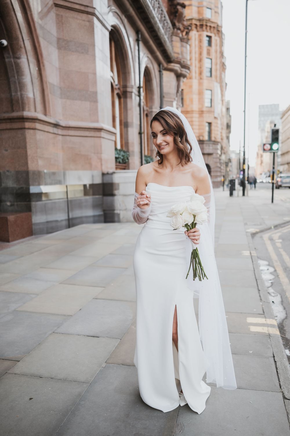 Bride in elegant off-shoulder white wedding dress holds small bouquet while standing on urban street near historic building.