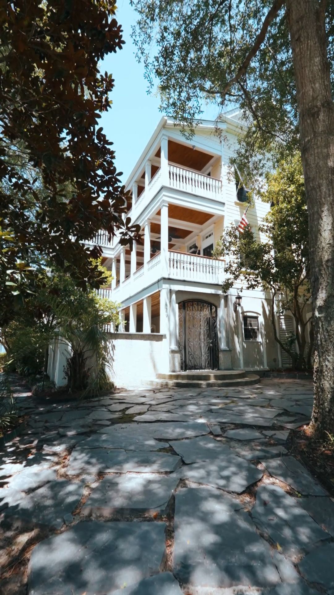 Elegant three-story white Southern mansion with double porches, arched entry, and stone pathway through mature trees.