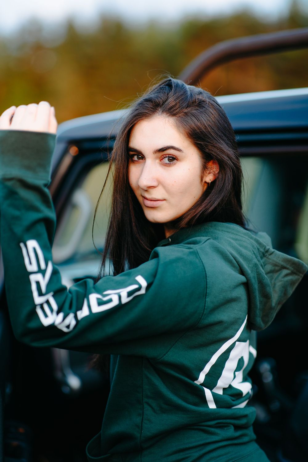 A person in a green jacket poses playfully while leaning out of a car window at sunset.