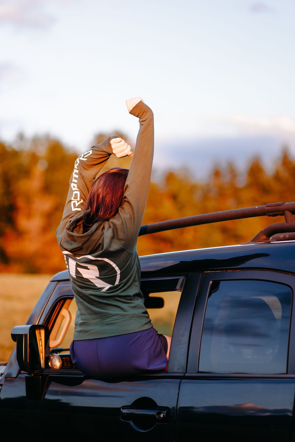 Someone sitting on top of a car with their arms raised against an autumn forest backdrop.