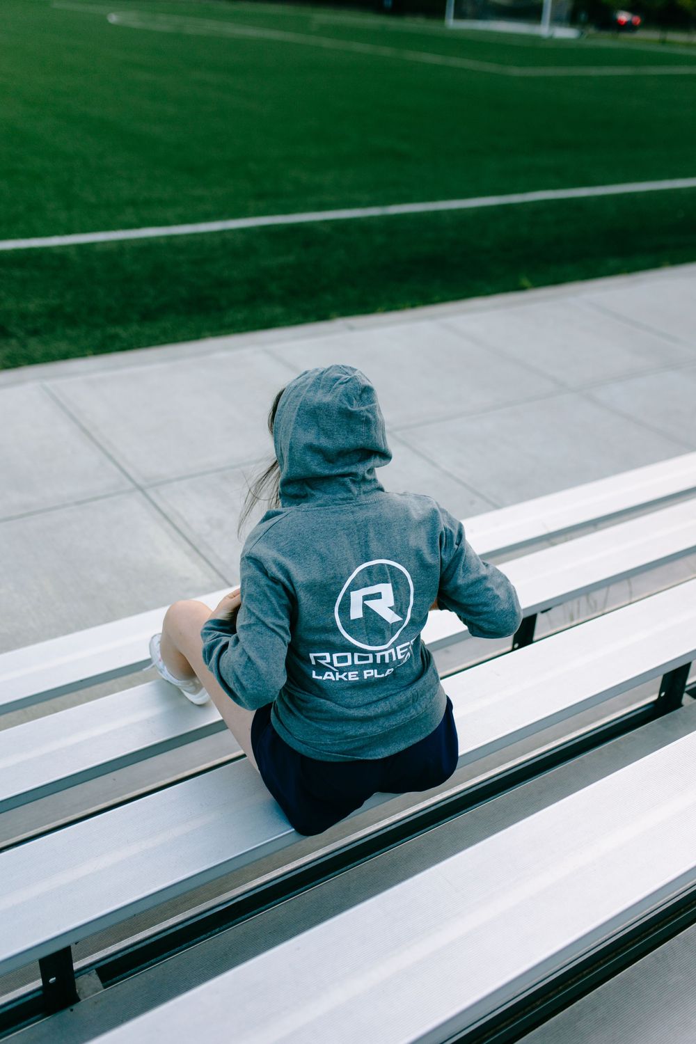 A person wearing a blue hoodie with a circular logo sits alone on metal bleachers overlooking a sports field.