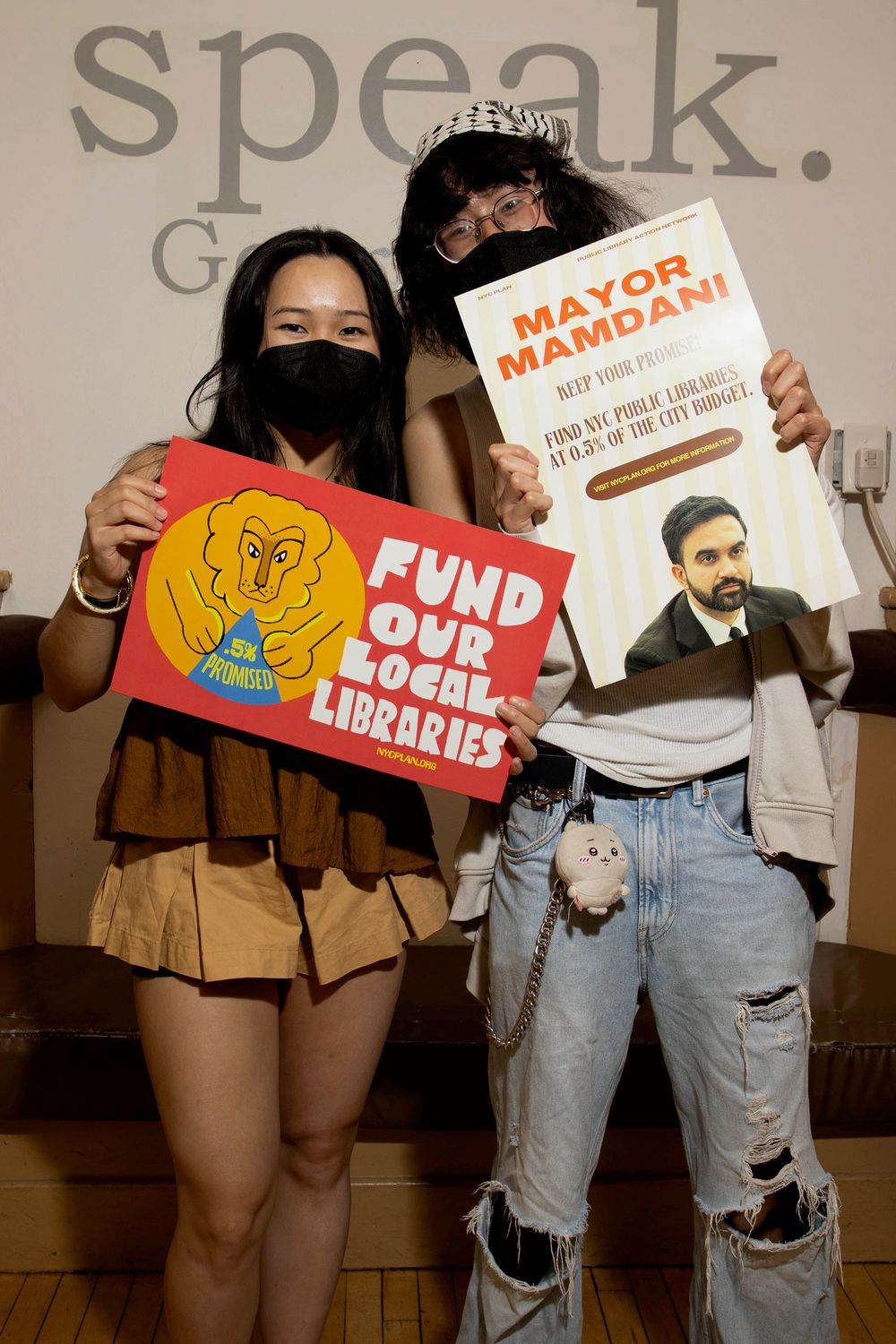 Two activists hold signs advocating to fund local libraries and urging Mayor Mamdani to protect the city budget.