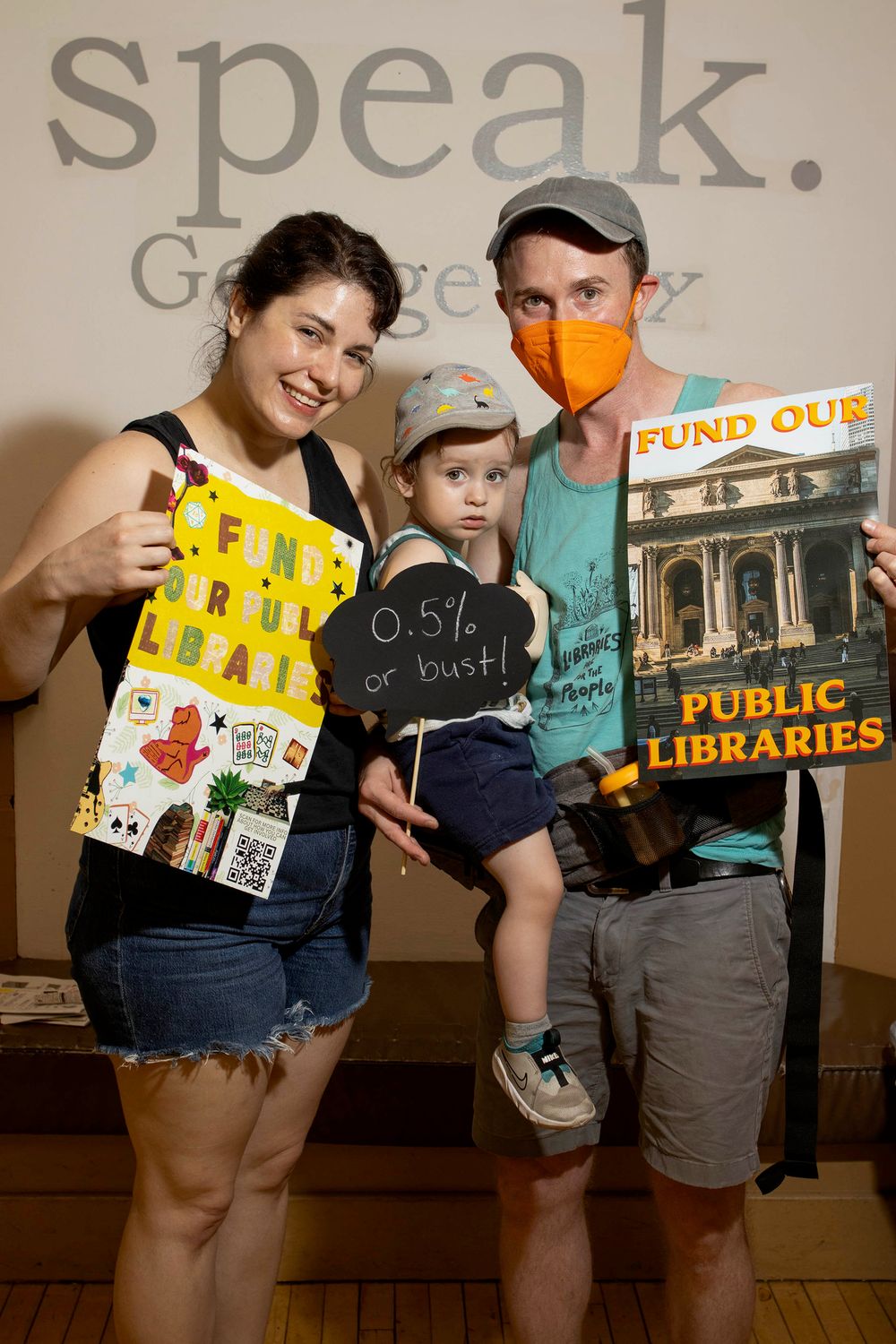 Family with toddler holds colorful signs supporting public library funding at a speak-out event.