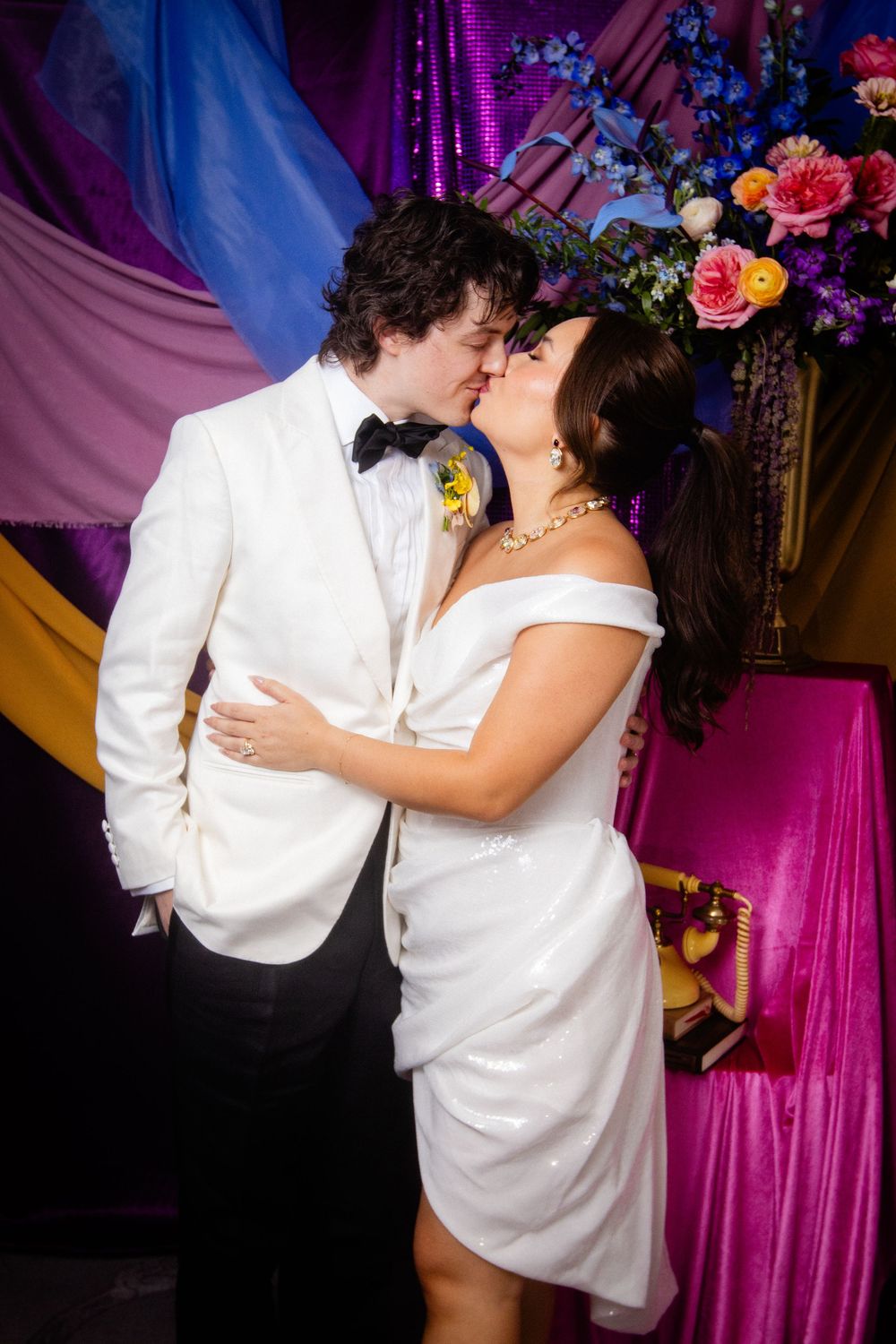 Couple dancing against colorful purple and blue draped backdrop with floral arrangements.