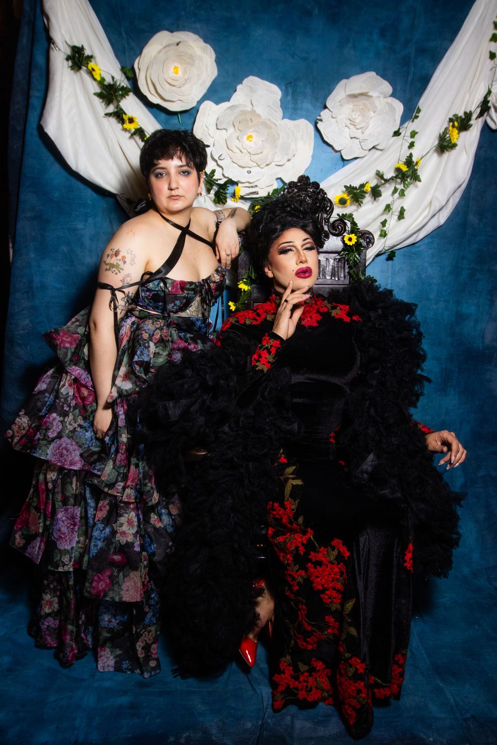 Dramatic portrait of two people in gothic formal wear against blue backdrop with paper flowers.