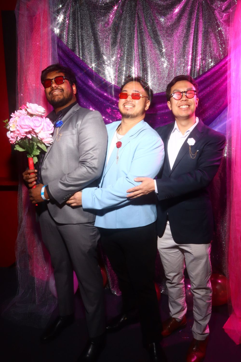 Three friends pose together against a sparkly backdrop with pink lighting, one holding pink flowers.