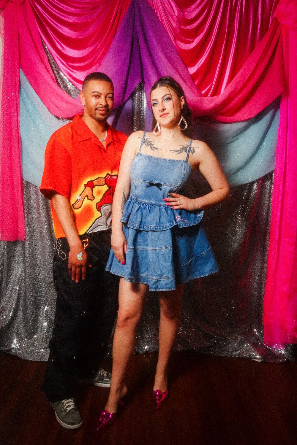 Two people posing in front of colorful draped backdrop, man in red shirt and woman in denim mini dress with pink heels.
