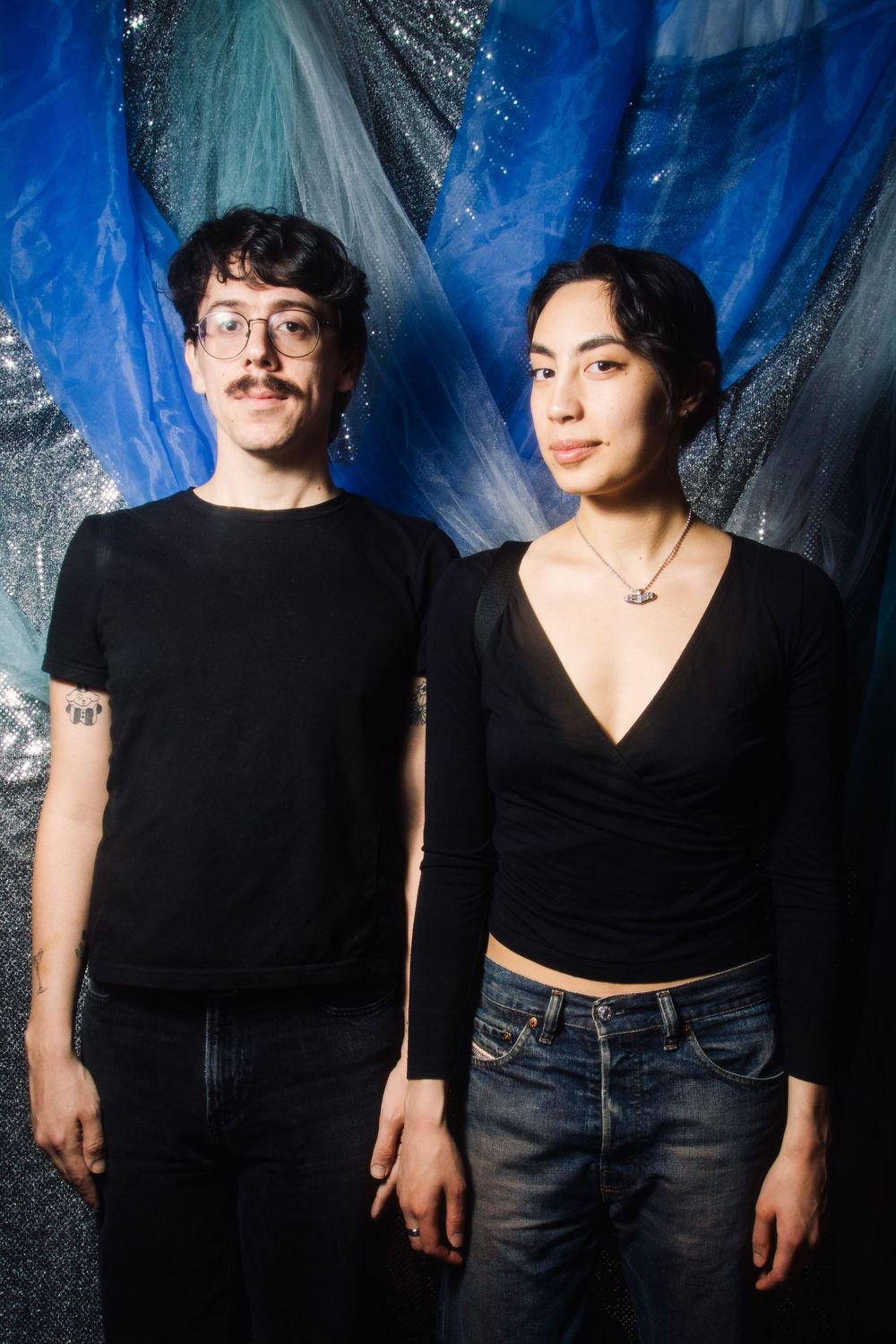 Two people in black outfits pose against a blue metallic backdrop, man with glasses and tattoo beside woman in wrap top.