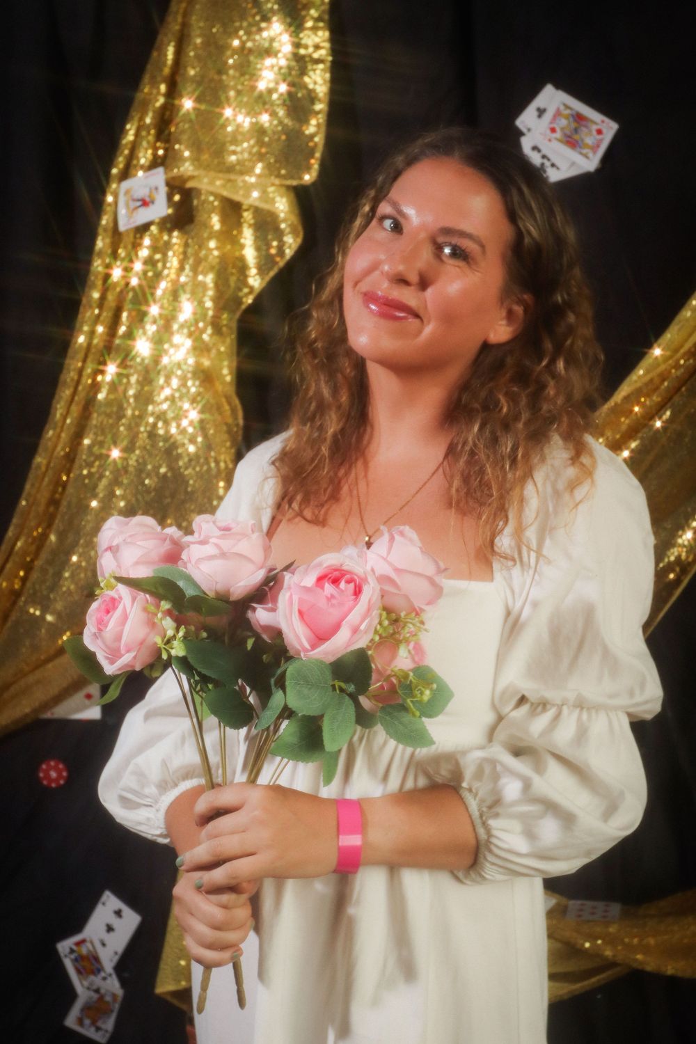 Woman in white dress holding pink roses with playing cards falling around her against a golden glittery backdrop.