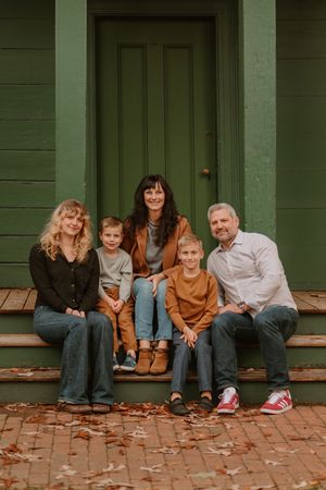 Family sitting on brick steps against green wall with autumn leaves during outdoor photoshoot.