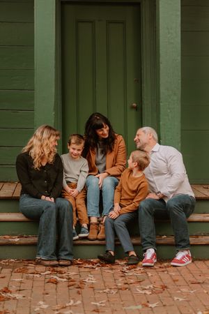 A family sits together on the steps of a green door, sharing a casual moment on an autumn day.