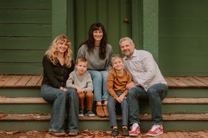 A family of five sits together on green wooden porch steps covered in autumn leaves.