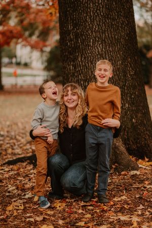 A tender autumn scene with fallen leaves beneath a large tree trunk shows a joyful family moment.