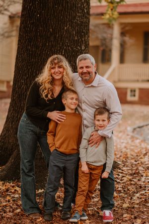 A candid autumn family photoshoot with fallen leaves surrounding their feet against a rustic building and tree backdrop.