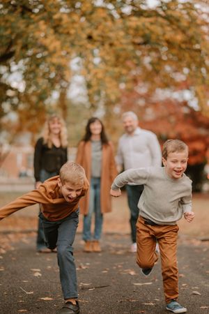 Two children run and play while adults stand in background surrounded by vibrant fall foliage.