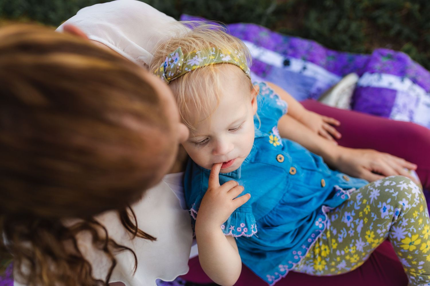 A small child in a blue romper lies on a purple blanket outdoors while being embraced.