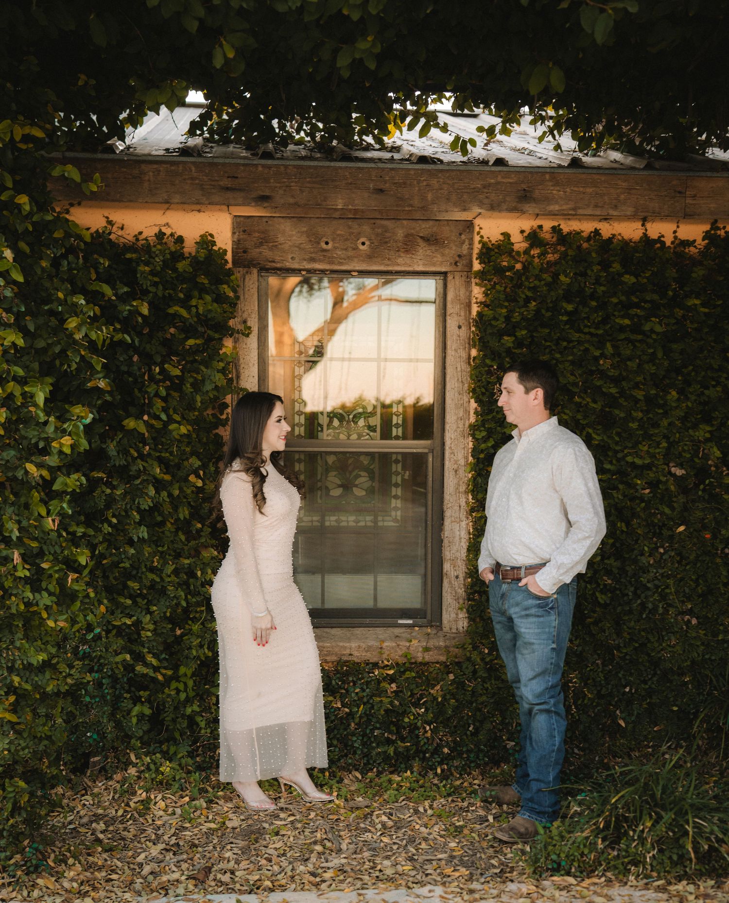 Couple in light colored outfits posing by rustic window with ivy covered walls.