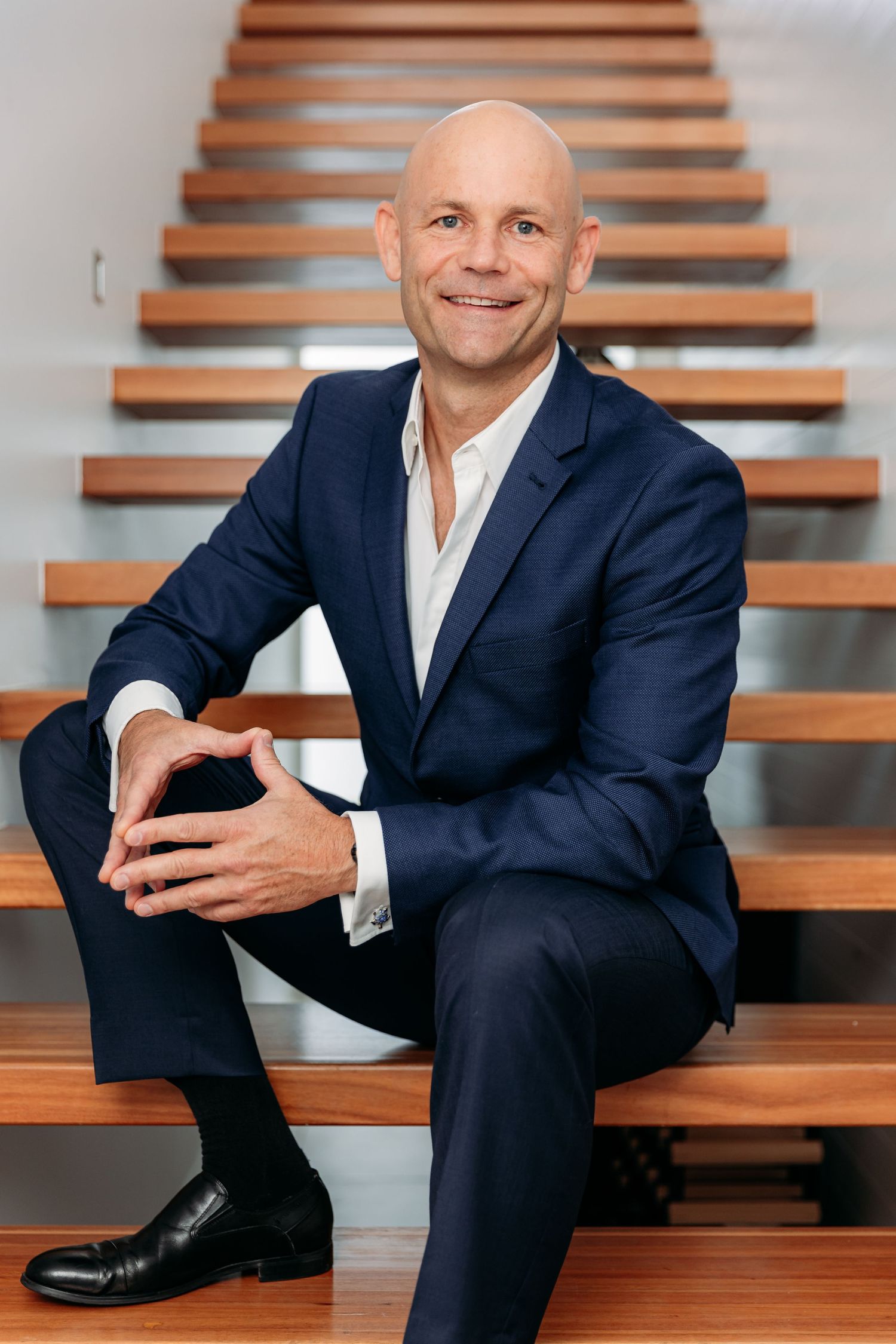 Professional in navy suit sits casually on modern wooden stairs in business portrait series.