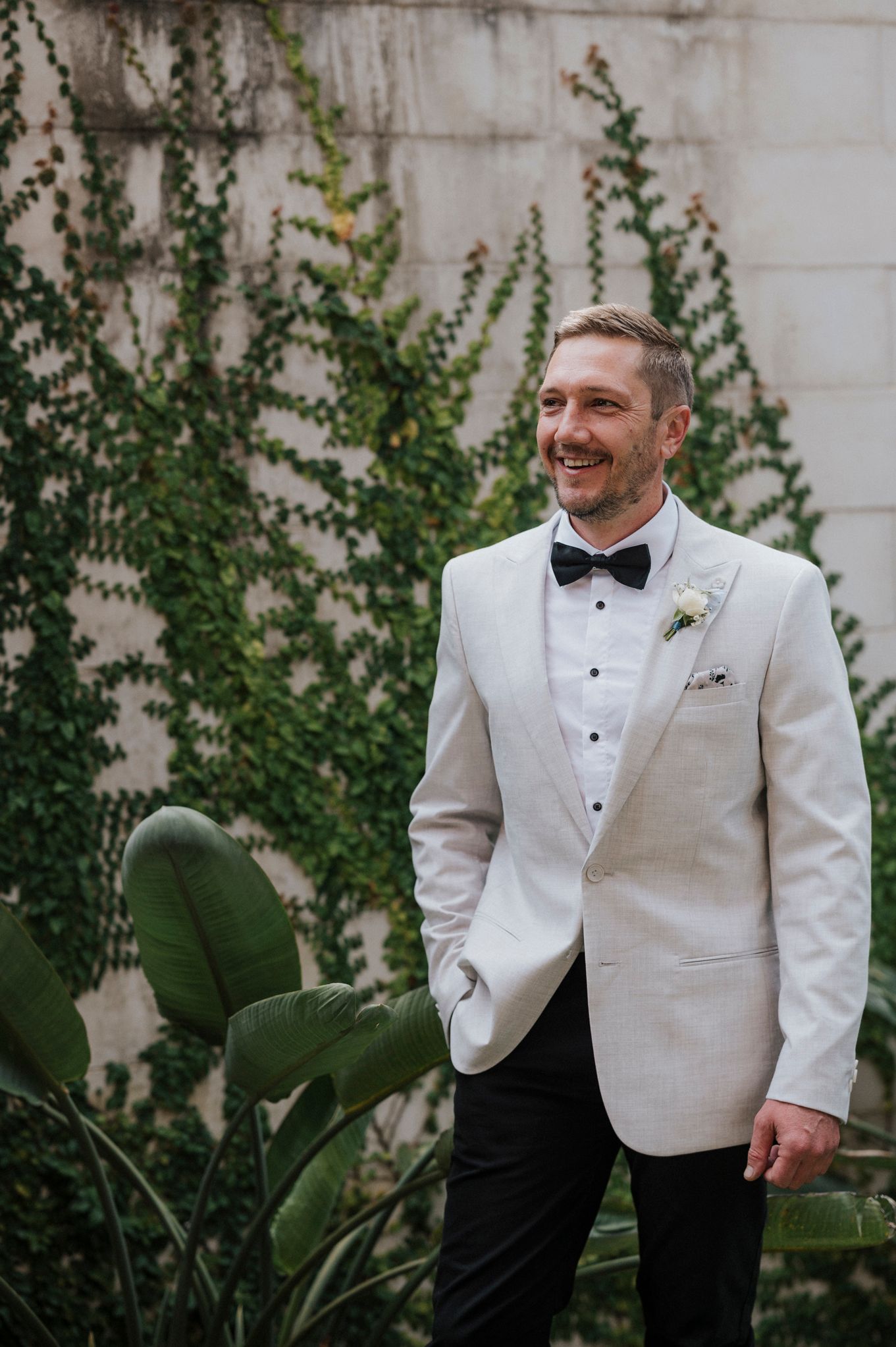 Wedding attire featuring white tuxedo jacket and black bow tie against vine-covered wall.