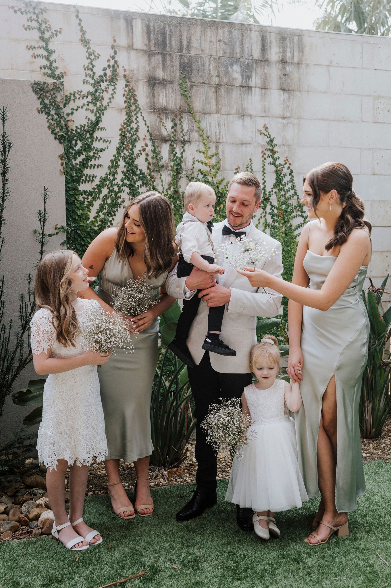 Wedding party poses playfully in garden setting with white wall and tropical plants.