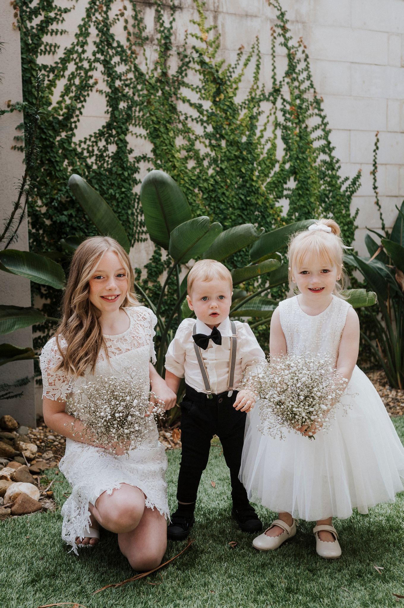 Children in formal white attire holding baby's breath bouquets pose on garden lawn.