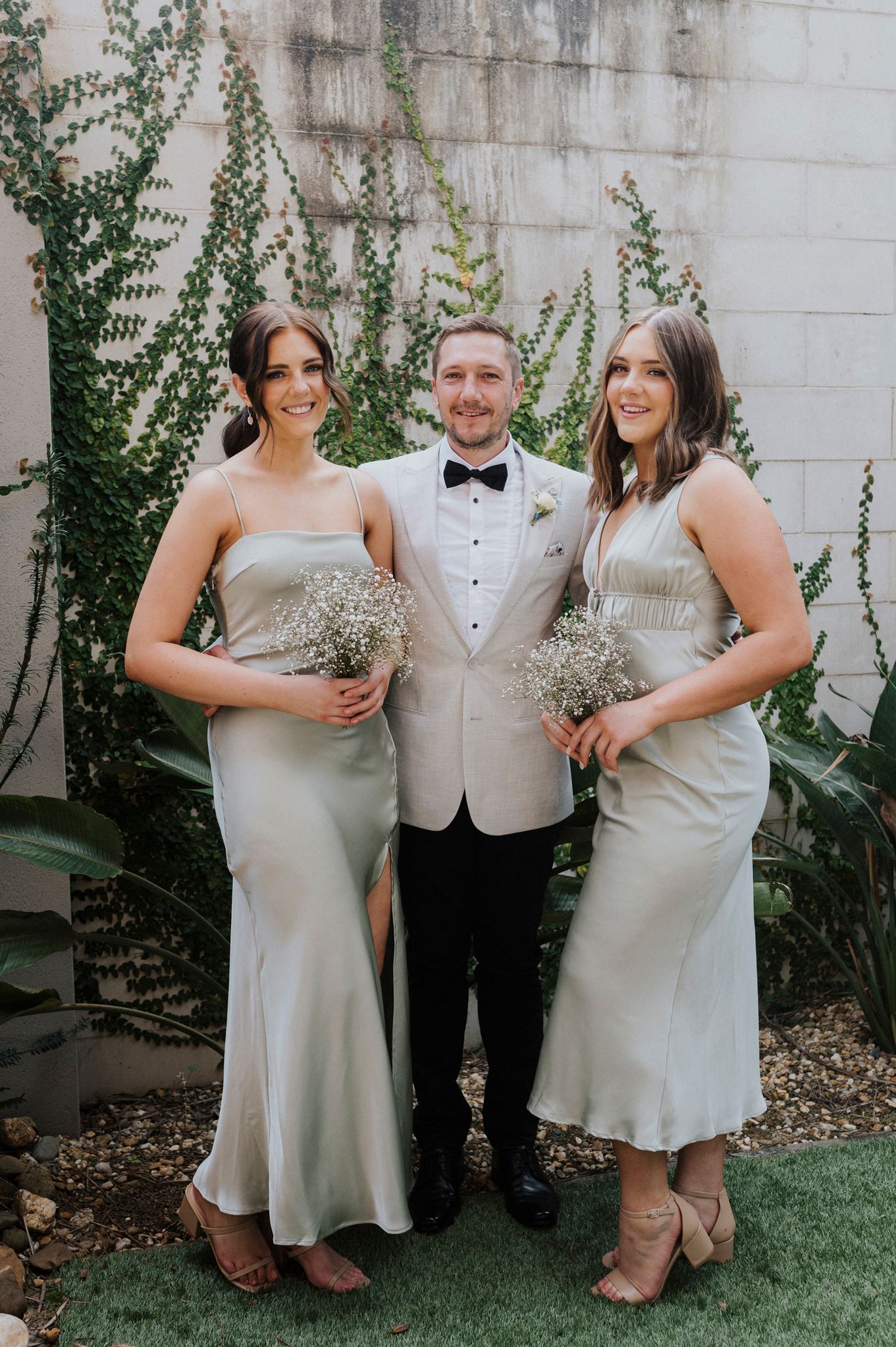 Wedding party in sage green dresses and tuxedo poses against white wall with climbing vines.
