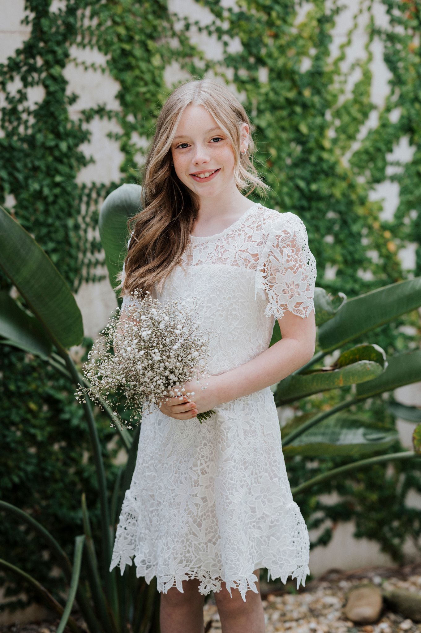 Young attendant in delicate white lace dress holding baby's breath flowers in garden setting.