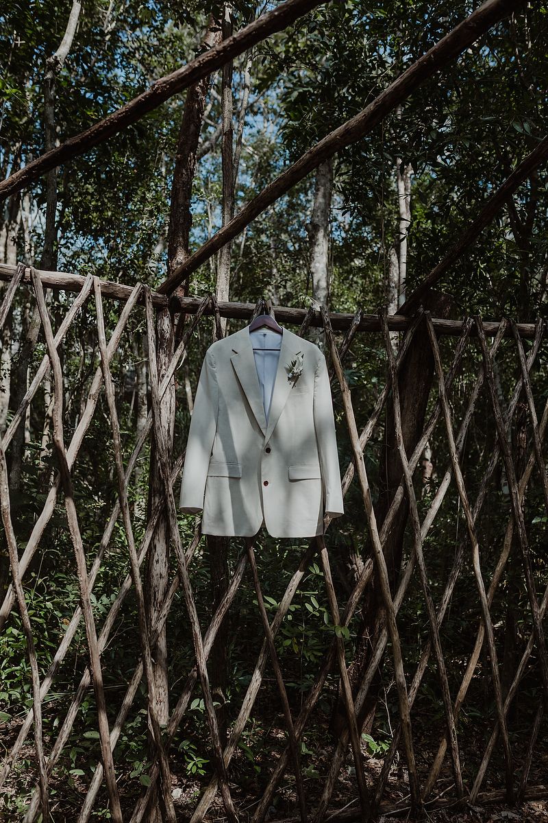 A white formal suit jacket hangs on a rustic wooden lattice fence against a leafy forest background.