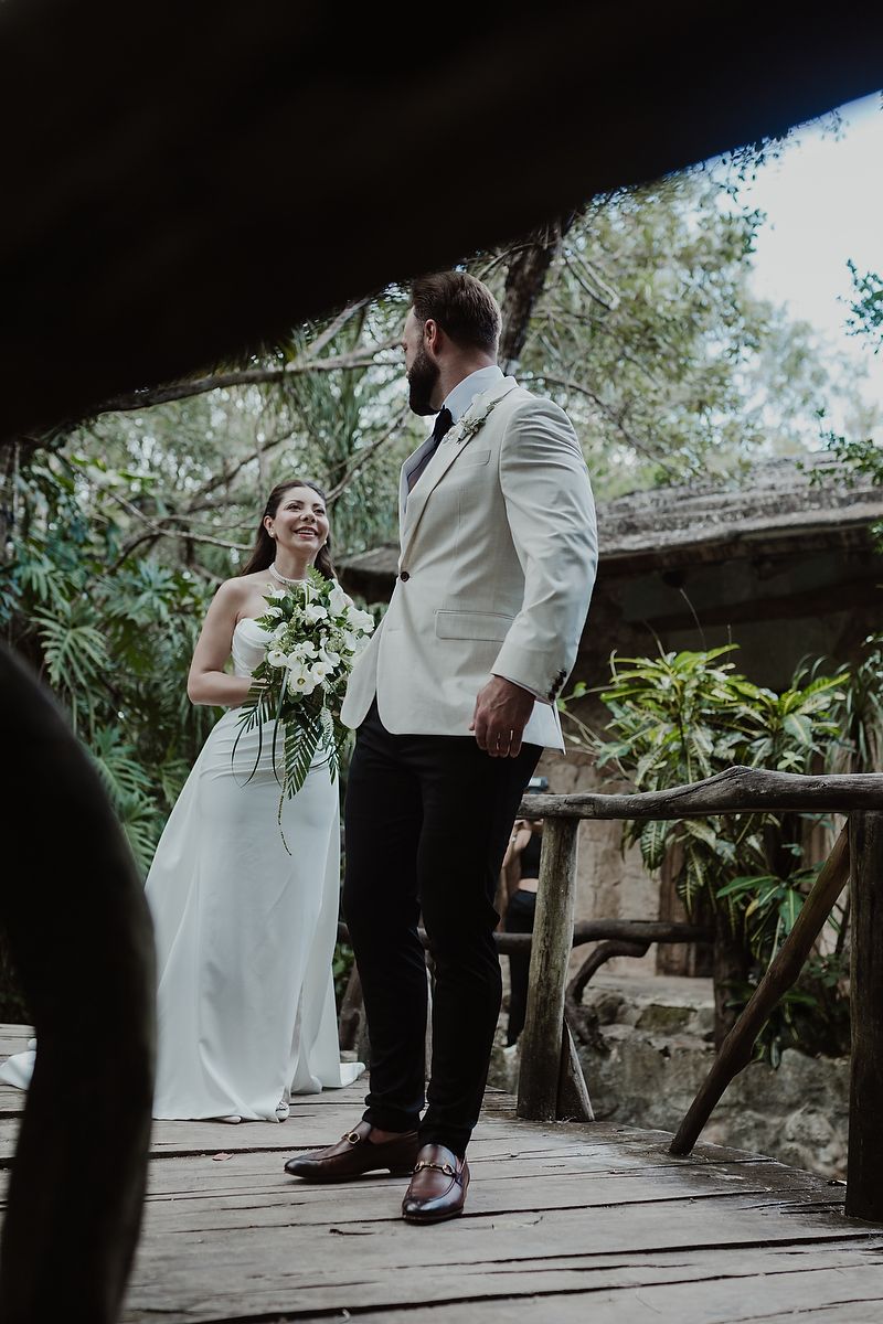 A wedding ceremony takes place on a wooden deck surrounded by lush greenery on a sunny day.