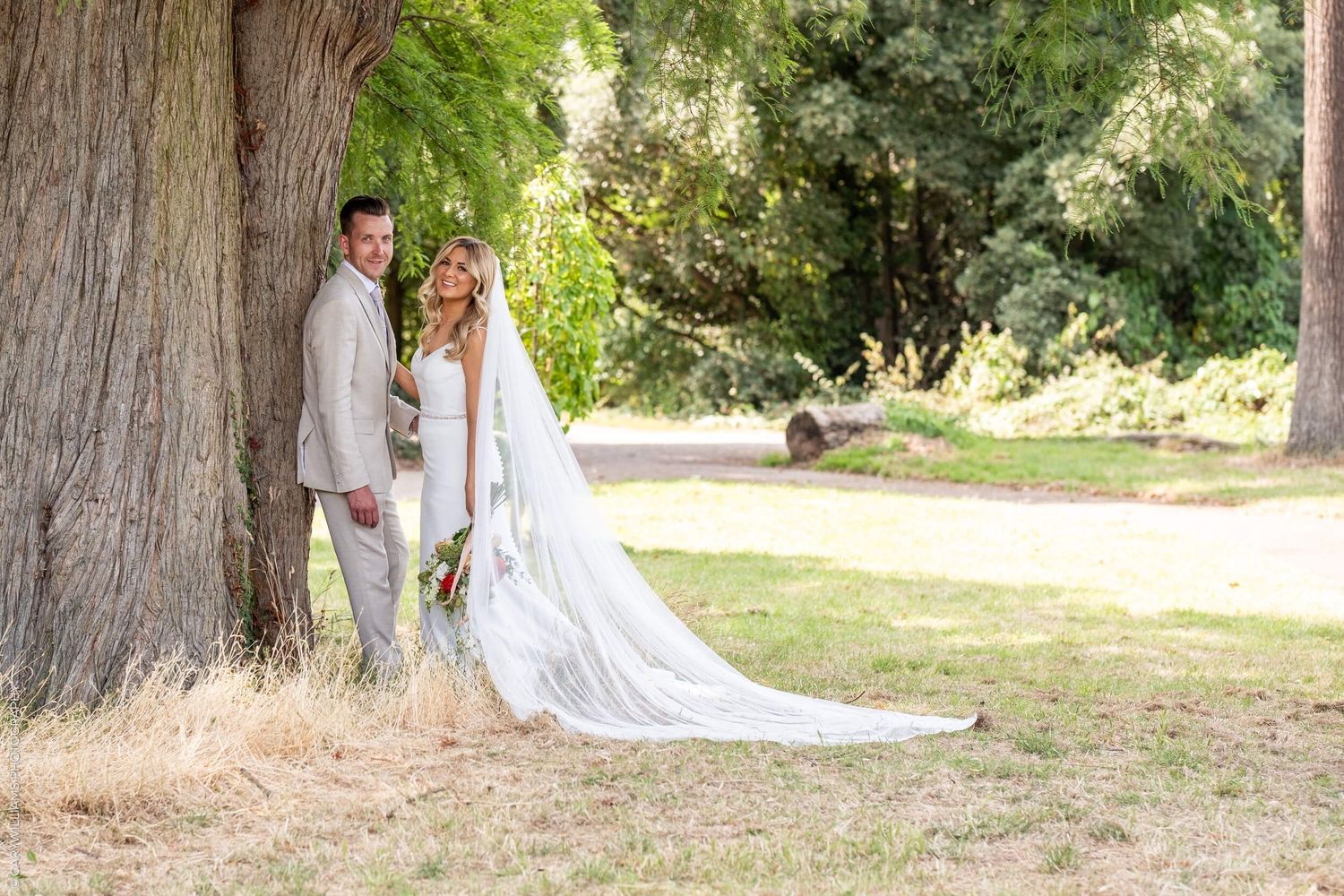 A couple poses together by a large tree trunk in a sunlit park setting, the bride wearing a flowing white gown with long train.