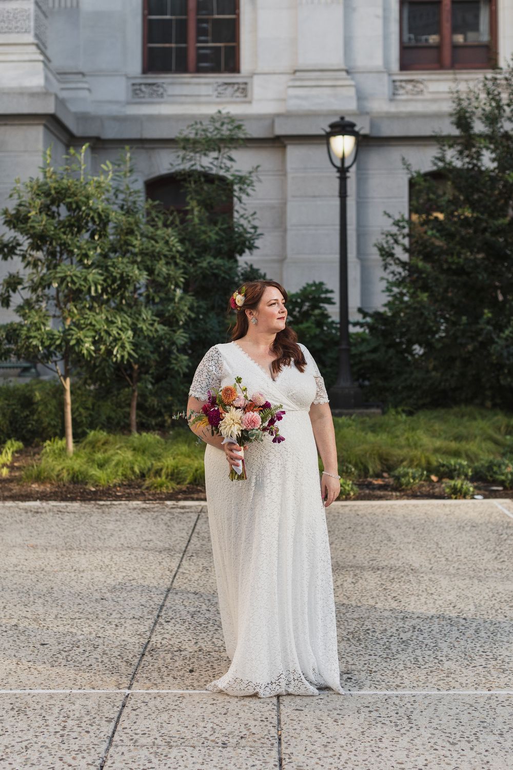 A person in a white dress holds a colorful bouquet on a gravel path near stone architecture at Philadelphia's City Hall.