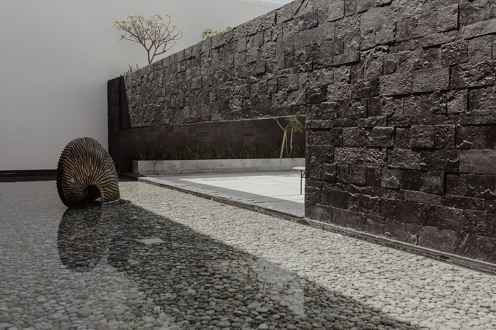 Modern stone wall garden feature with pebble pathway, water element, and decorative woven sculpture in minimalist outdoor space.