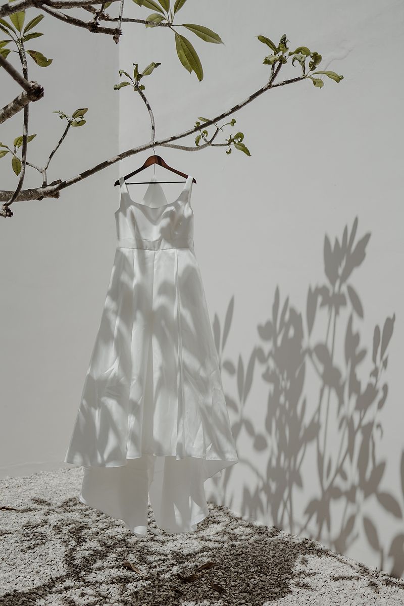 White satin wedding dress hanging on wooden hanger from tree branch, casting floral shadows on white wall in sunlight.