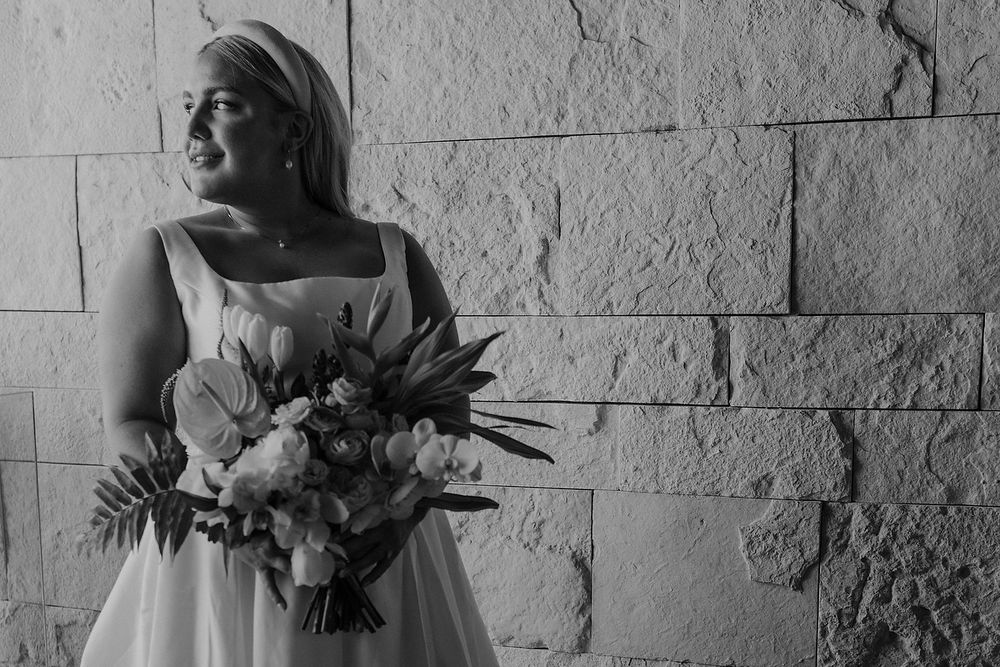 Black and white photo of a bride holding a bouquet of tropical flowers, standing against a stone wall.