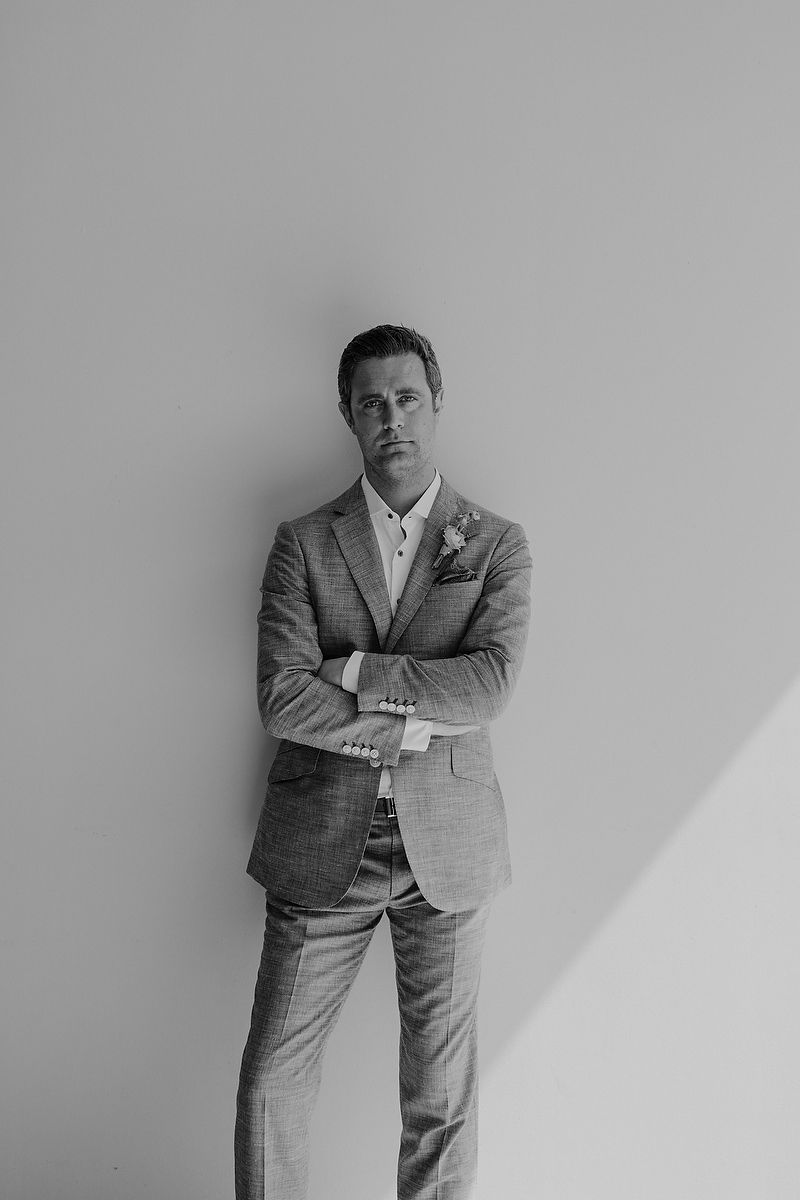 A groom in a tailored suit with boutonniere stands arms crossed against a wall in a black and white portrait.
