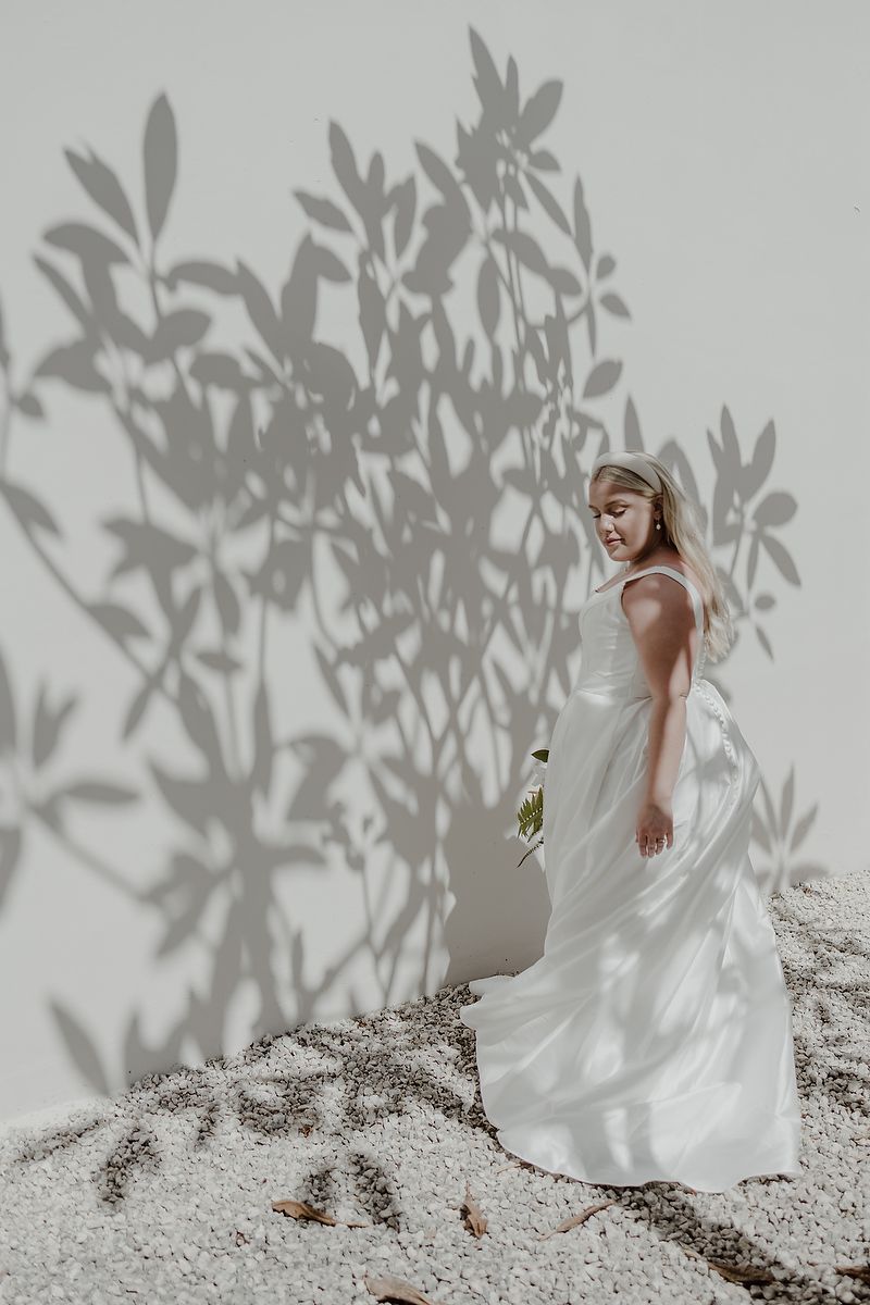 A bride in a white gown stands on sand with plant shadows cast on a white wall behind her.
