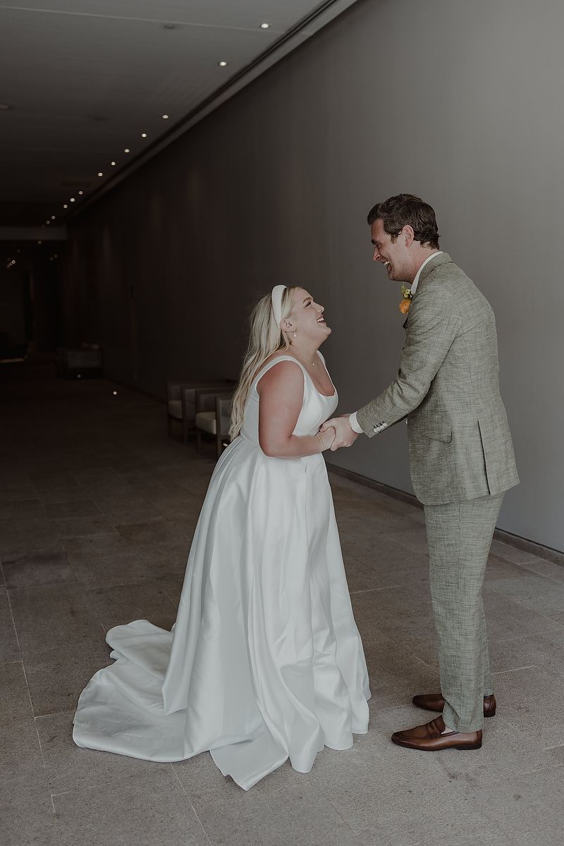 Bride in white gown and groom in gray suit hold hands and share a joyful moment in a modern indoor hallway.