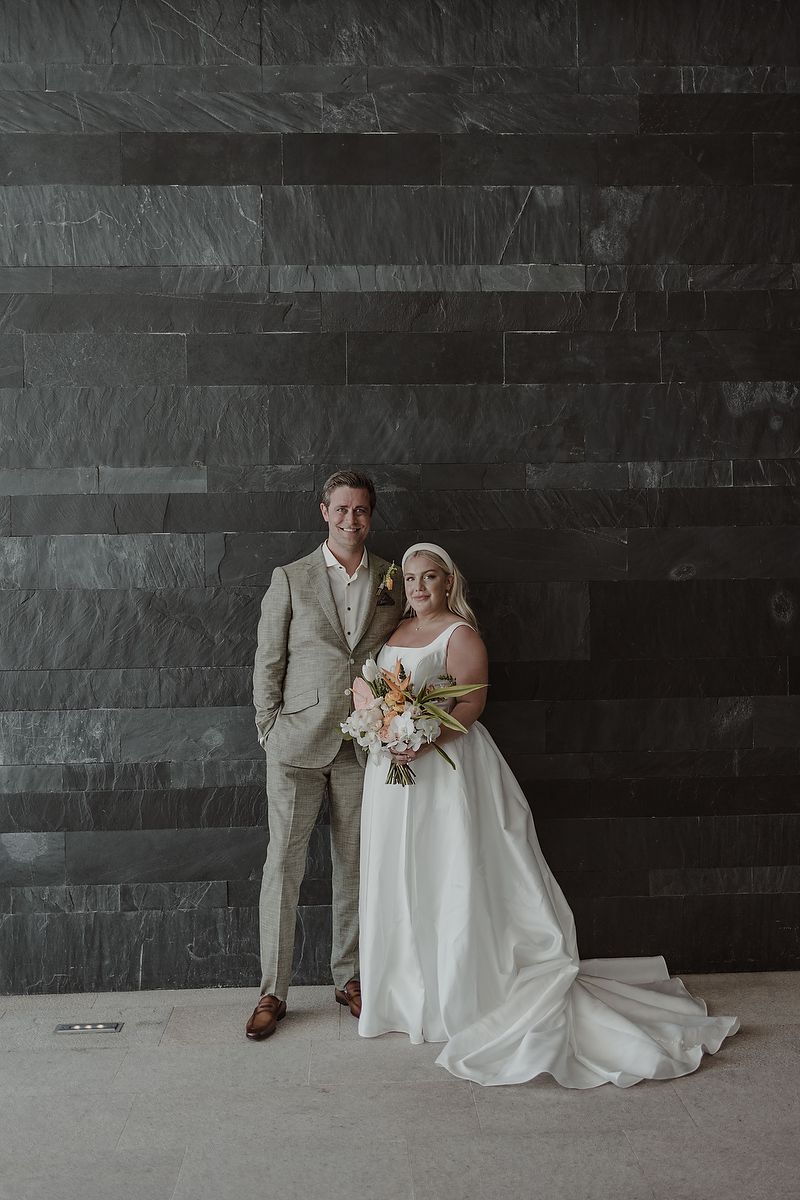 A smiling bride and groom pose together in front of a dark slate tile wall, bride holding bouquet.