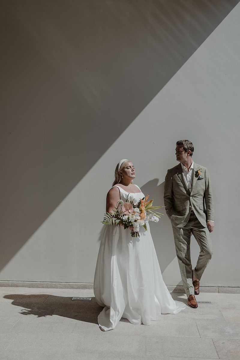Bride in white gown holding tropical bouquet stands with groom in olive suit against minimalist wall in natural light.