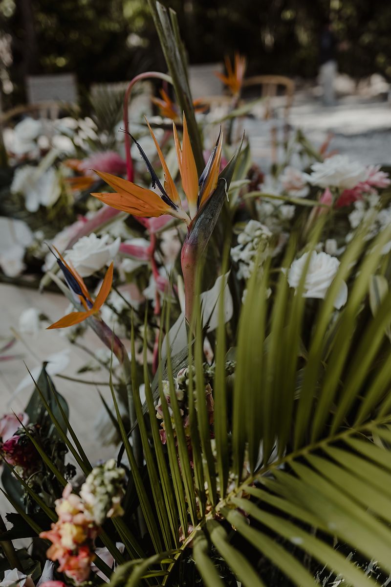 Tropical bird of paradise flowers and palm leaves in a lush floral arrangement at an outdoor wedding reception.