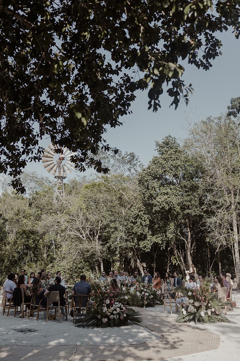 Outdoor wedding ceremony surrounded by lush trees with floral arrangements and a windmill backdrop on a sunny day.