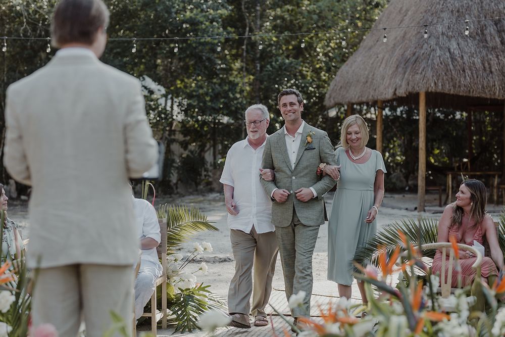 Groom walks down outdoor wedding aisle escorted by elderly parents at tropical venue with thatched roof.